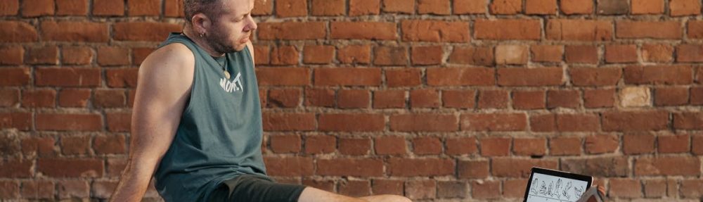 Athletic man sitting and resting on a mat in an urban gym, with a tablet and weights visible.