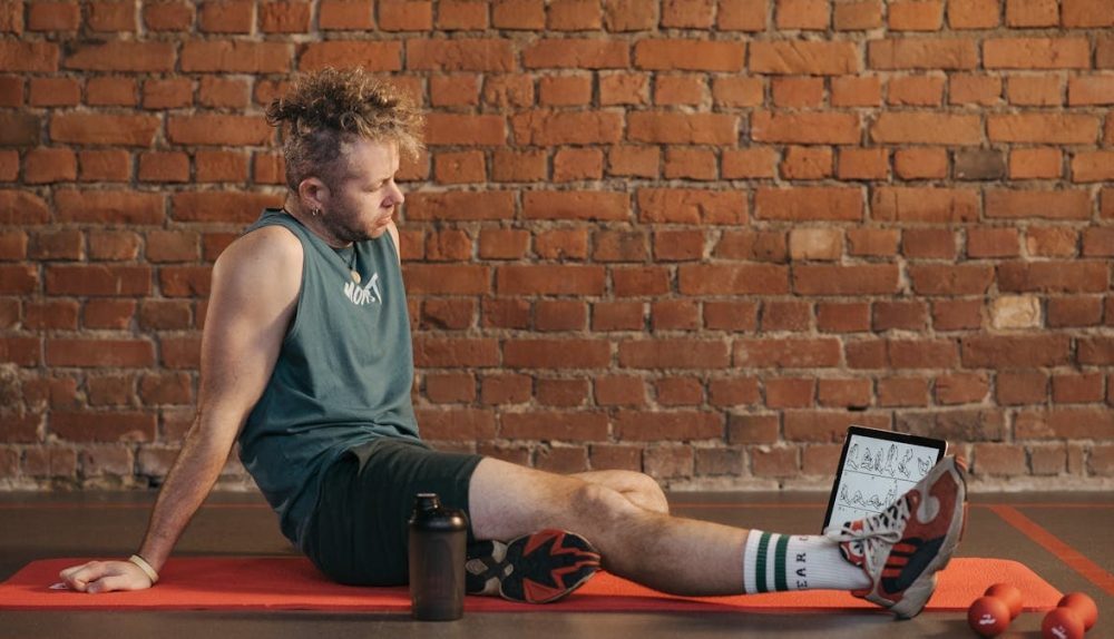 Athletic man sitting and resting on a mat in an urban gym, with a tablet and weights visible.